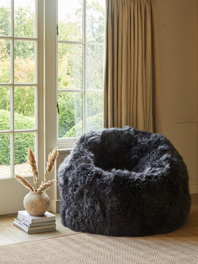 Fluffy dark gray bean bag chair in a room with large windows and beige curtains.